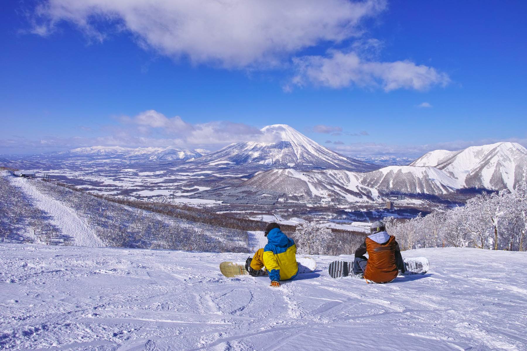 Two snowboarders resting on a groomed slope above a snow-covered valley, Mount Yotei in the distance