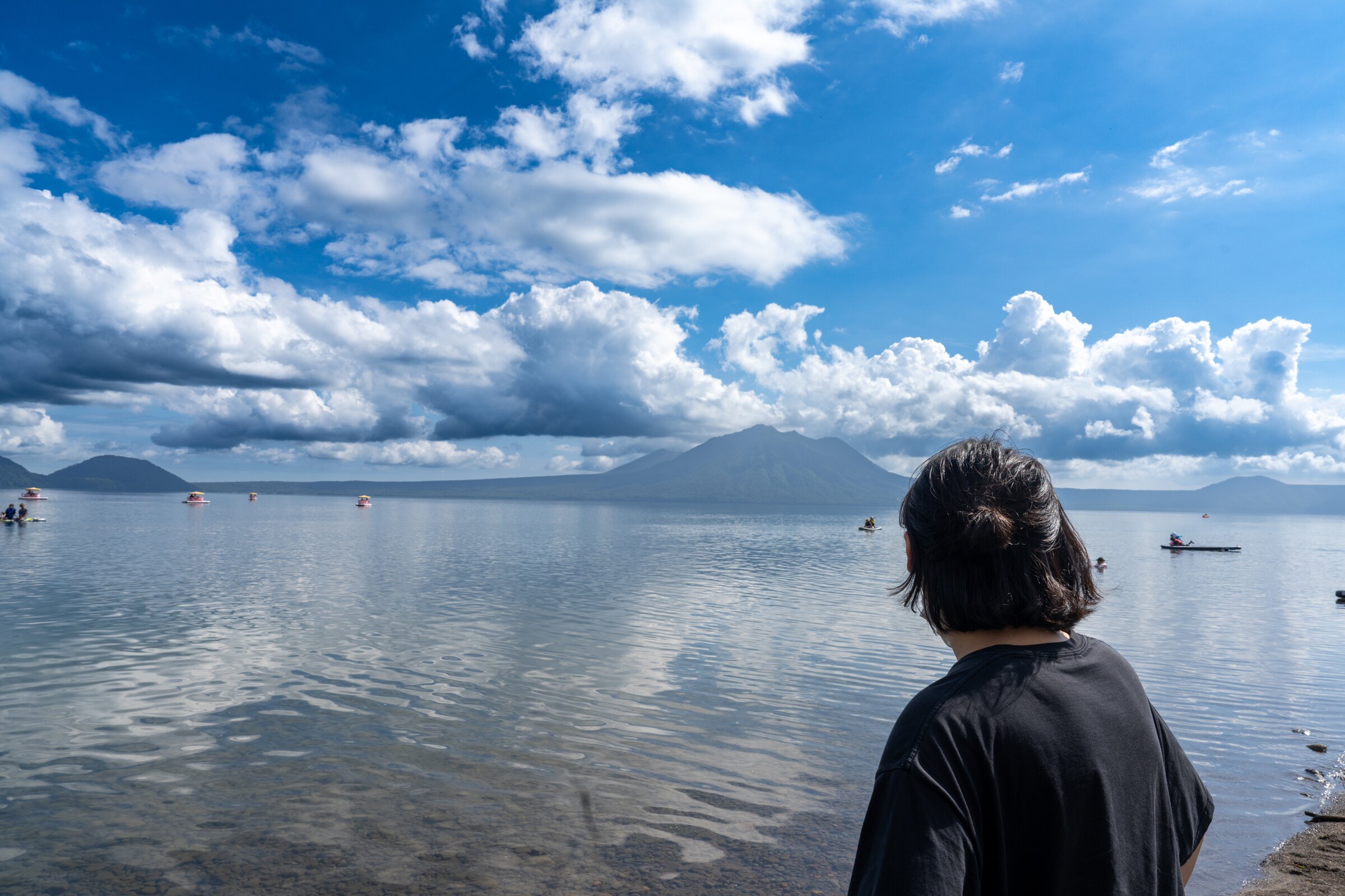 Traveler looking across a calm Hokkaido lake toward green mountains beneath summer clouds